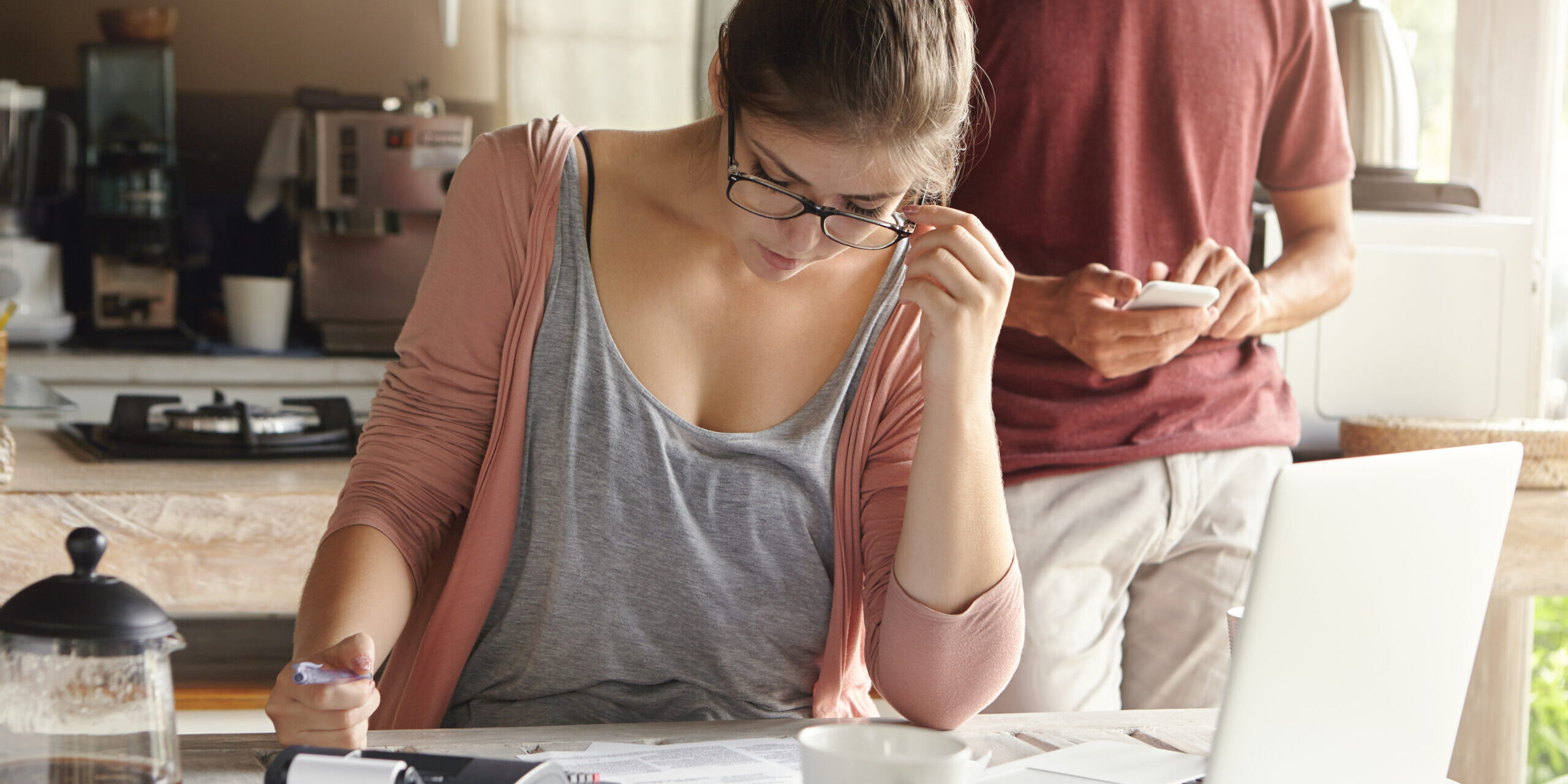 Serious young woman with hair bun wearing glasses looking through finances, planning family budget, sitting at kitchen table in front of open laptop, her husband using mobile phone in background
