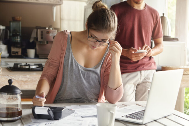 Serious young woman with hair bun wearing glasses looking through finances, planning family budget, sitting at kitchen table in front of open laptop, her husband using mobile phone in background